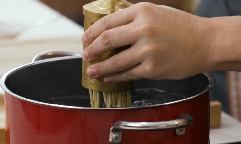 Hand pressing noodles through brass tool into a red pot of boiling water.